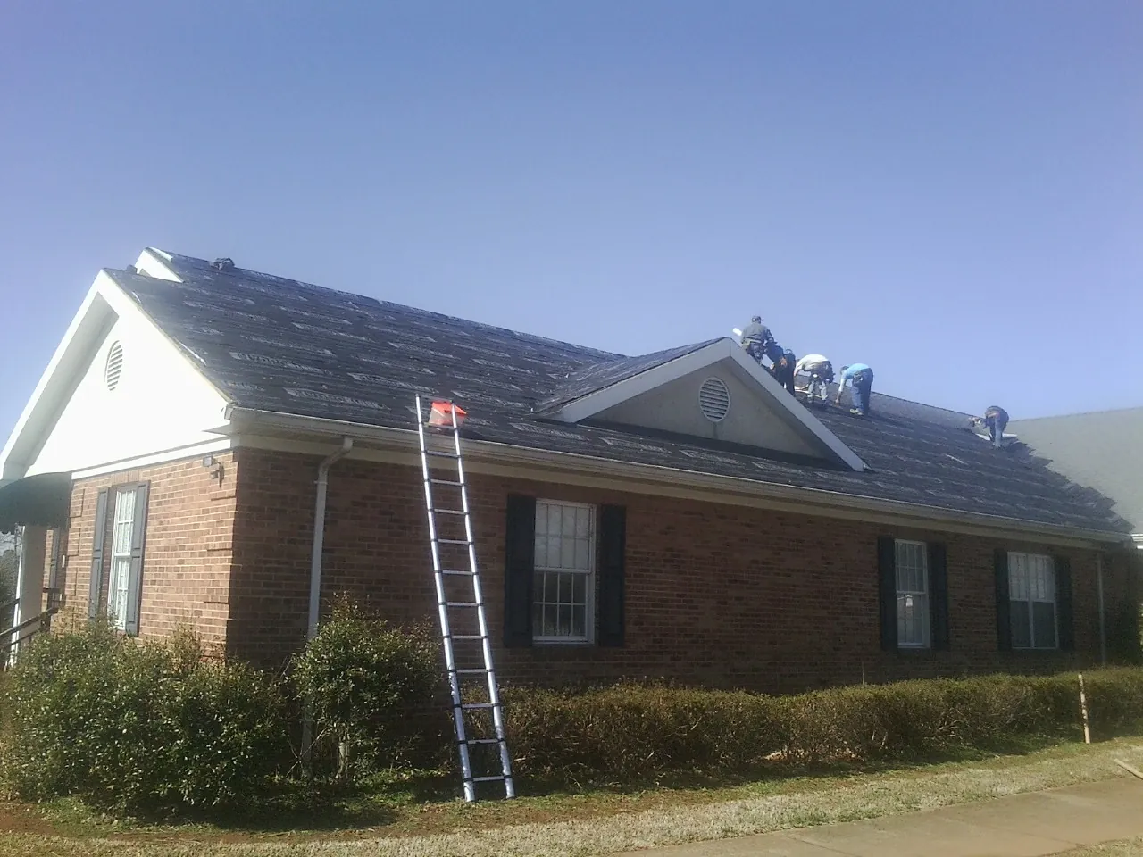 Crew tearing shingles off a brick ranch roof with a ladder set — Metal Roof Repair work by our crew in Arona