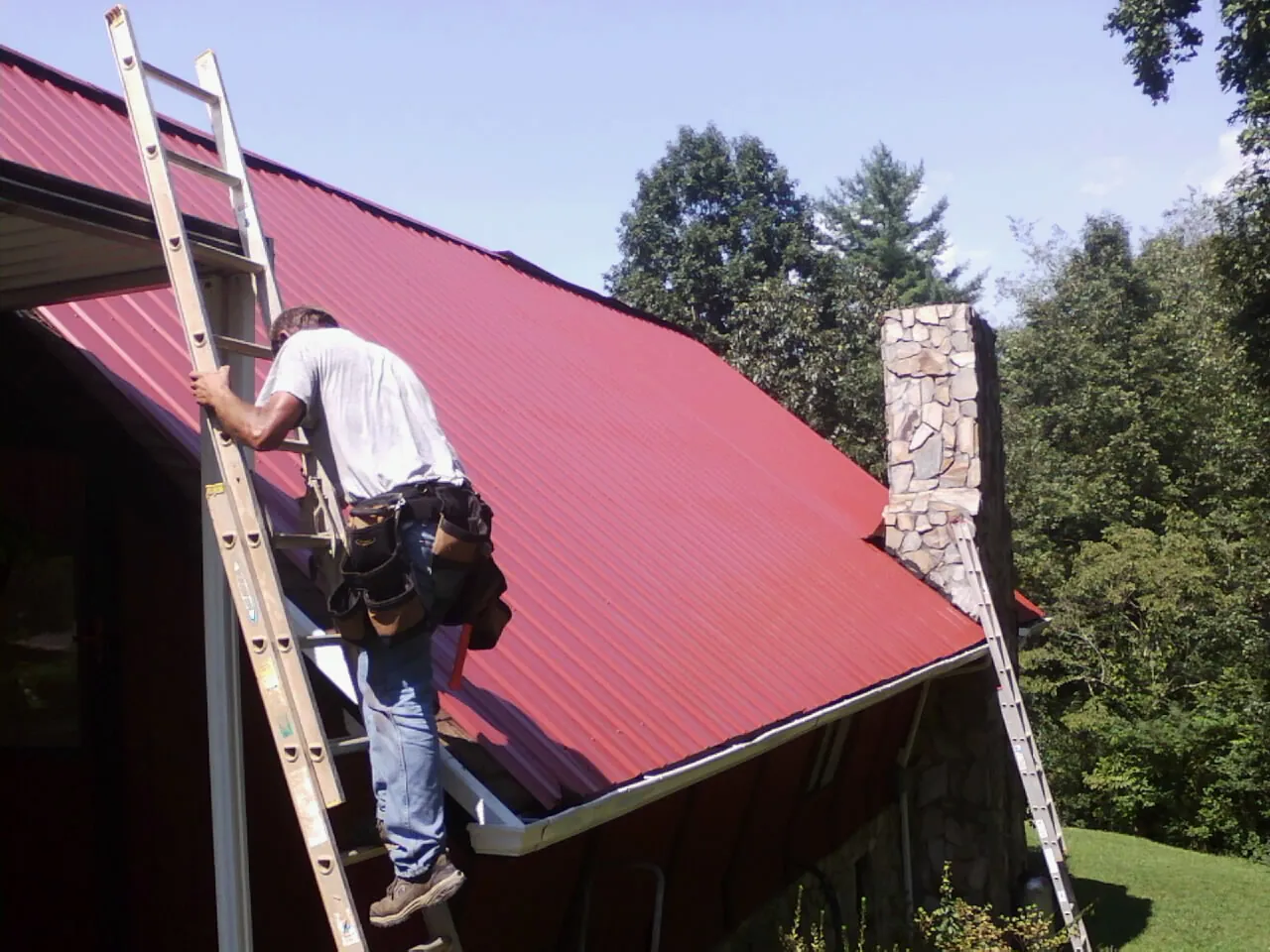 Roofer on a ladder beside a red metal roof and stone chimney — Soffit & Fascia Repair by Roofing Brothers in Arona